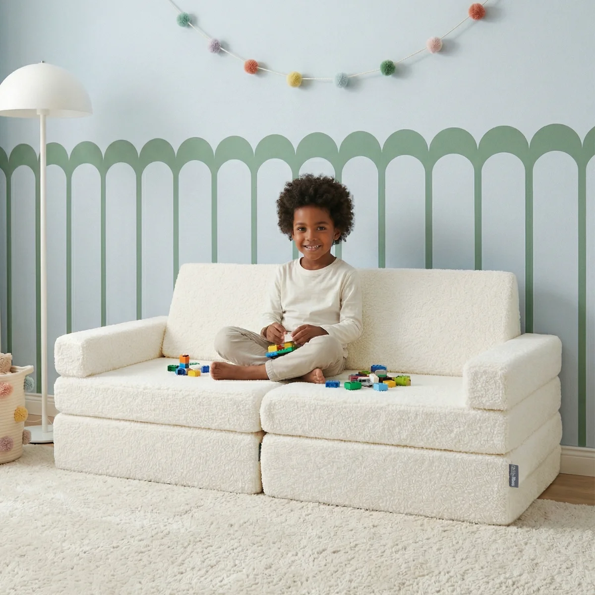 Kind sitzt auf einem cremefarbenen Teddy-Spielsofa in einem zarten Pastellraum